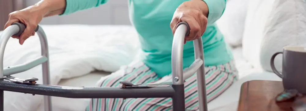 An elderly woman sitting on a bed and leaning on her walker, about to stand up